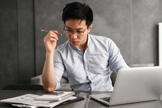 Portrait of a concentrated young asian man using laptop computer while sitting at the table with paperwork at home